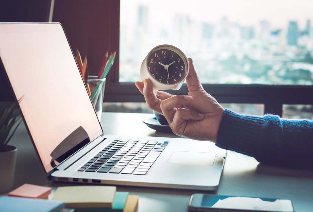 a person looks at an analogue clock while using their laptop and phone.