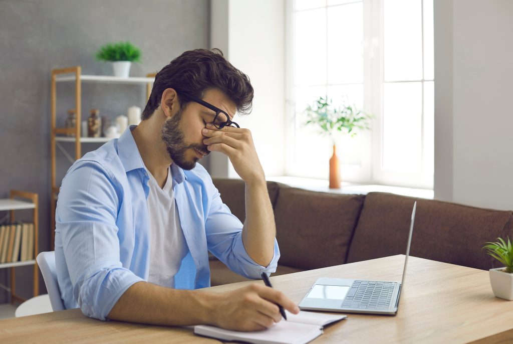 A man rubs his tired, strained eyes while working at a desk.