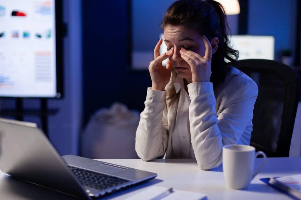 A young woman rubs her tired eyes while working at her desk at night.