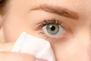 Close-up of a person’s green eye with long lashes, gently wiping the under-eye area using a white cotton pad.